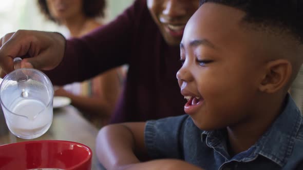 African american father smiling while pouring milk in cereal bowl on his son at home