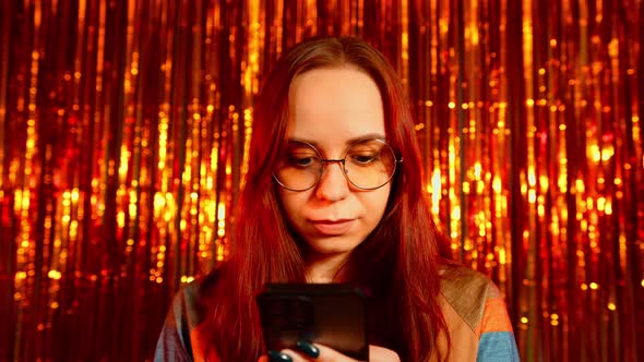 Young Woman in Glasses Browsing Mobile Phone on Background of Shiny Tinsel