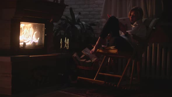 A Girl is Sitting in a Rocking Chair and Reading a Book Near the Fireplace