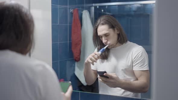 Longhaired Handsome Man Looking at His Phone Whilst Brushing His Teeth  Middle Shot