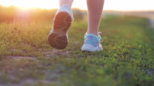 A Child in Bright Sneakers Walks on the Green Grass in the Meadow During Sunset