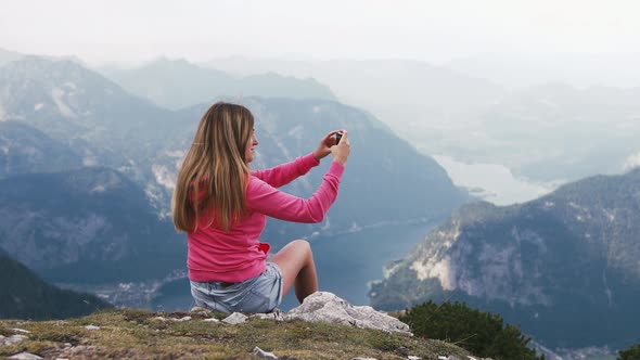 Young Woman Taking Photo with Smart Phone at Mountain Peak Wth Stunning Mountain Lake Background