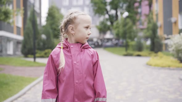 Portrait of Cute Little Girl in Waterproof Coat Walking Along the Alley Outdoors. Pretty Caucasian