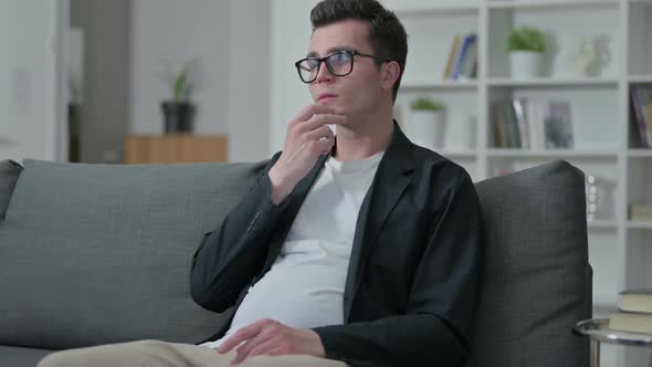 Pensive Young Male Designer Sitting on Sofa and Thinking at Home