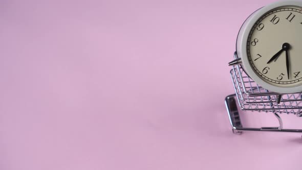 Hand rolls a decorative supermarket trolley with a white alarm clock inside on a pink background 