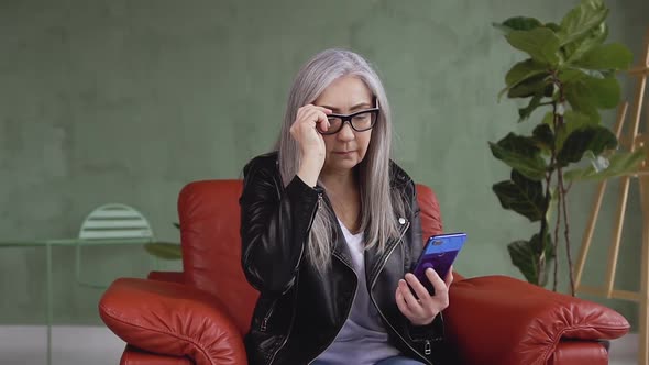 Modern 60-Aged Woman in Glasses which Using Her Smartphone while Sitting in Comfortable Chair