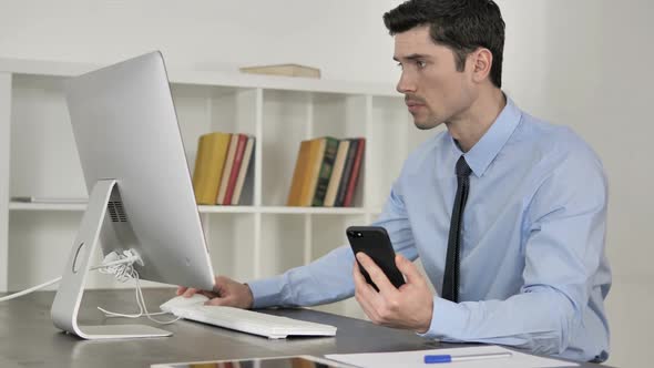 Businessman Using Smartphone While Working on Computer