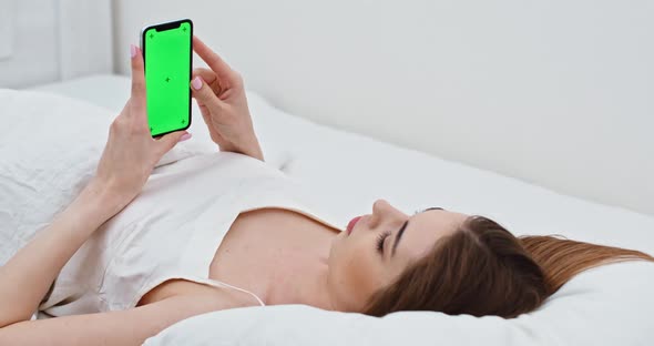 Young Woman Lying in Bed and Watches a Vertical Video on Her Smartphone, Green Screen, Girl Is