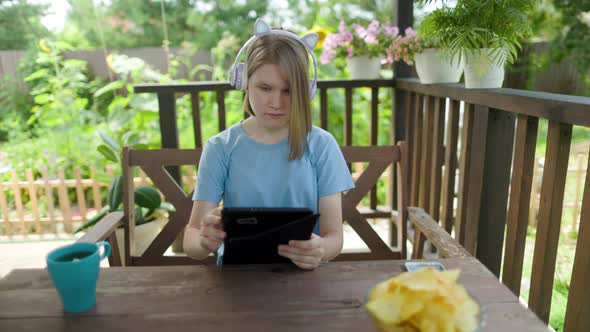 Teenage Girl Sits At Wooden Table In a Summer Cafe In Headphones With Phone