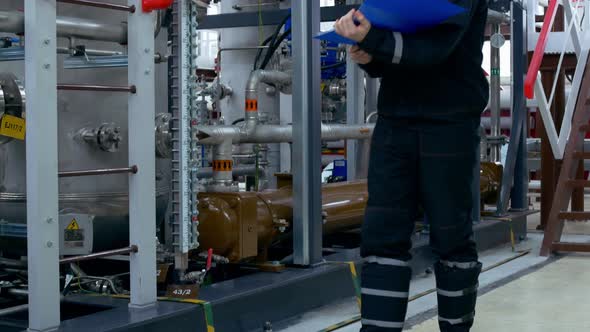 A Factory Worker in a Helmet and Overalls Inspects the Compressor in the Shop