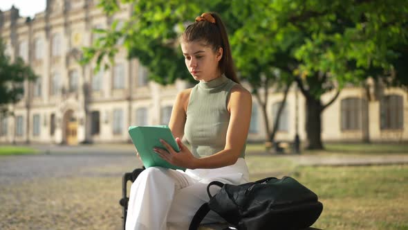 Young Thoughtful Woman Surfing Internet on Tablet Sitting on Bench on Sunny University Yard Looking