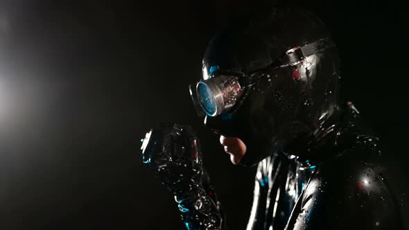 a Woman in a Black Latex Suit Helmet and Glasses Touches Neck and Opens Palms Against a Dark