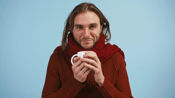 Young Man is Inhaling a Flavor of Tea or Hot Medicine in Cup and Smiling Posing on Blue Background