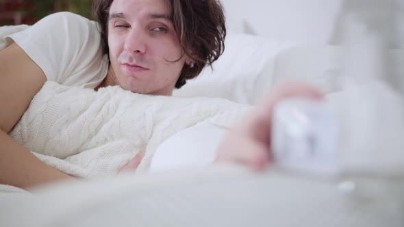 Closeup Portrait of Sleepy Young Man Looking at Alarm Clock and Waking Up with Shocked Facial