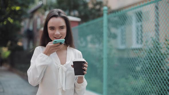 Beautiful Girl with Brown Hair Holding a Coffee Paper Cup and Using Her Smartphone Voice Commander
