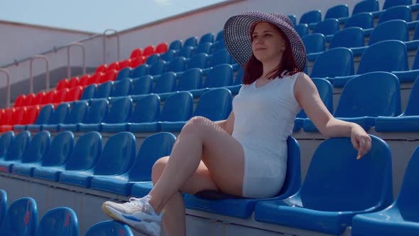 Young woman in in white dress and elegant hat looks away, sitting on stadium bleachers alone.