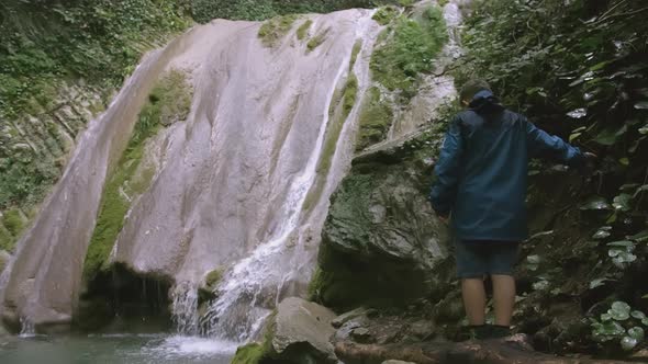 Guy in a Blue Windbreaker is Standing on a Hiking Trail and Looking at a Waterfall