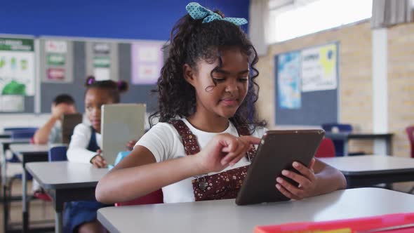 Portrait of happy mixed race schoolgirl sitting at classroom, using tablet, looking at camera