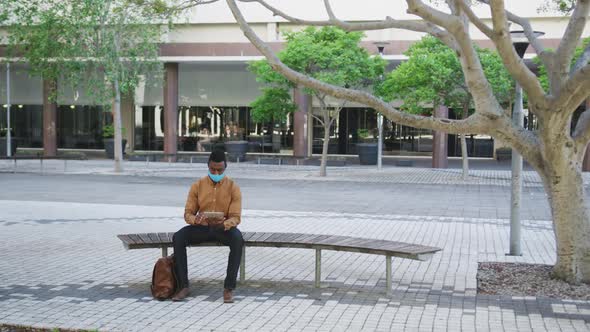 African american businessman wearing face mask using tablet sitting on bench