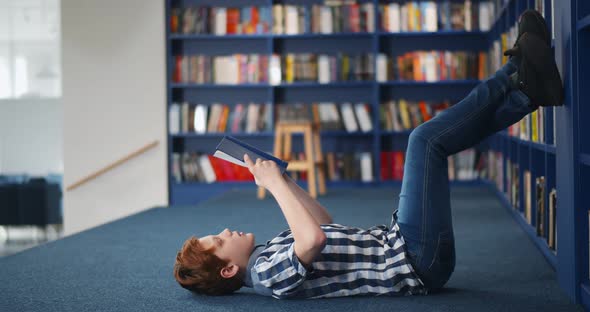 Redhead Preteen Boy Lying on Floor and Reading Book in Library
