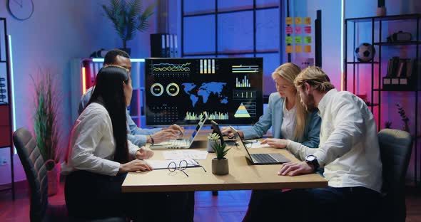 Young People Sitting Around Workplace in Modern Evening Office and Browsing Information on Computers