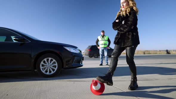 Upset Beautiful Slim Woman Kicking Road Cone with Blurred Auto Instructor Standing at Background