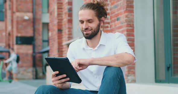 Man in White Shirt with Beard Sitting on Stairs and Working on Computer Tablet