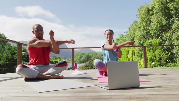 Happy biracial couple on terrace exercising, stretching with online lessons