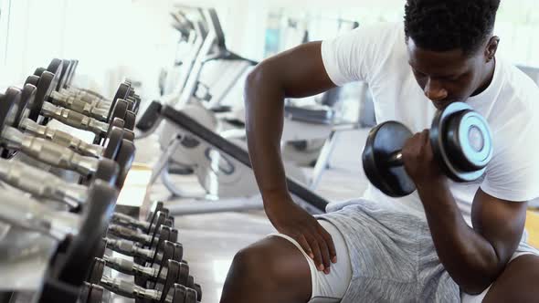 Young African American Man Sitting and Lifting a Dumbbell Close to the Rack at Gym