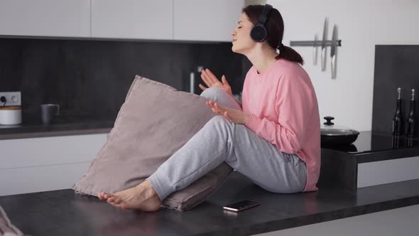 A Cheerful Young Woman Sitting on Kitchen Counter in New Home Listening to the Music