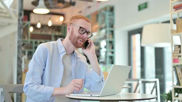 Redhead Man with Laptop Talking on Smartphone in Cafe 