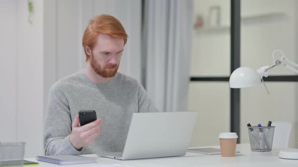 Beard Redhead Man with Laptop Checking Smartphone at Work