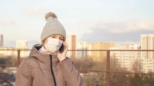 Young Woman in a Medical Mask on the Street with a Smartphone in Her Hand Against the Background of