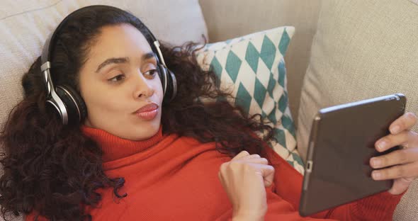 Happy woman lying on sofa using tablet enjoying music on her headphones