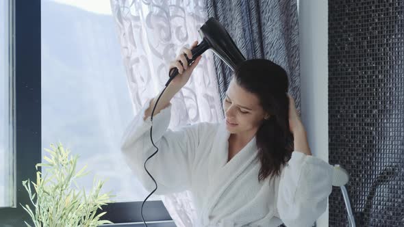 Charming brunette woman in bathrobe drying hair with blow dryer after taking shower