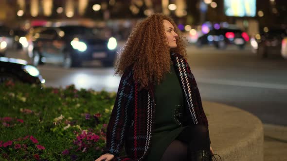 Portrait of a Beautiful Young Woman Resting on a Bench in the Center of the City in the Background