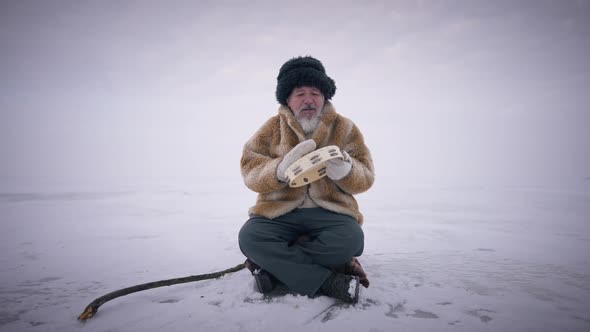 Wide Shot Old Mongol Sitting on Frozen Snow Outdoors Playing Tambourine in Slow Motion