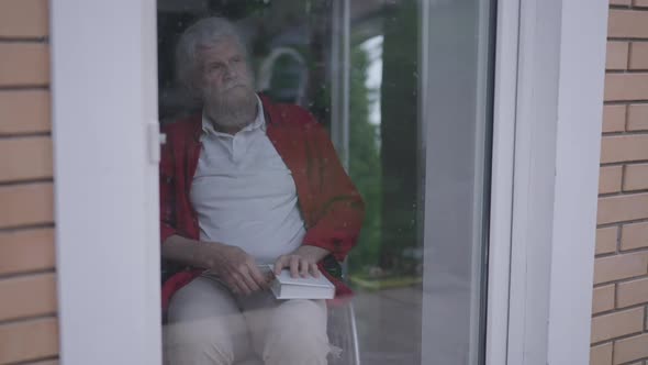 Depressed Thoughtful Senior Handicapped Man Closing Book Looking Out Window on Glass Door