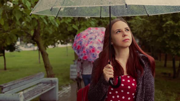 Young Melancholic Beautiful Woman with Transparent Umbrella Standing in Alley in Rain