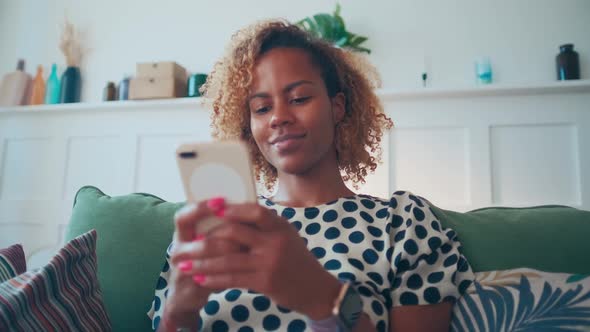 Young African American Woman Smiling Playing Mobile Phone Apps Sits on Sofa