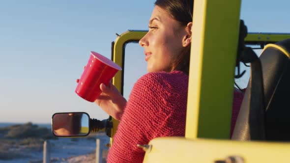 Happy caucasian woman sitting in beach buggy by the sea drinking