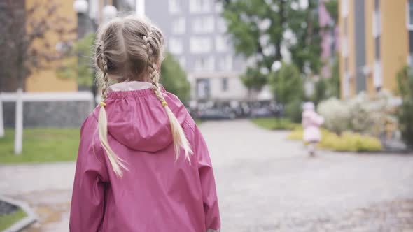 Back View of Little Girl with Pigtails in Pink Waterproof Coat Walking Outdoors. Blond Pretty