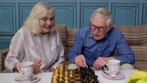 Retired Senior Couple Talking Drinking Tea Playing Chess in Modern Living Home Room Lounge Together