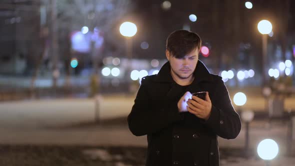 A man against the background of night lights is typing text on a smartphone .