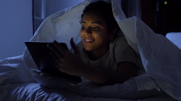 Woman with Tablet Pc Under Blanket in Bed at Night