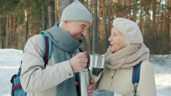 Happy Senior Couple Clinking Cups Drinking Hot Tea From Thermos and Chatting Outdoors in Winter Park