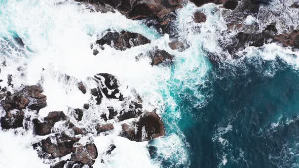 Aerial View of Waves Break on Rocks of Faroe Islands Cliffs in a Blue Ocean