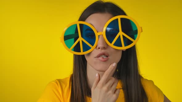 Woman in Funny Yellow Hippie Sunglasses is Posing on the Yellow Background