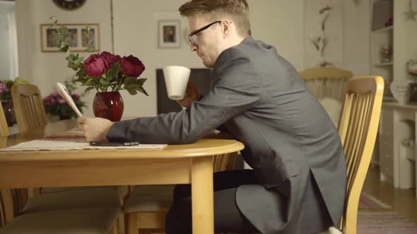 A young business man sits at a table wearing a suit and reads the newspaper