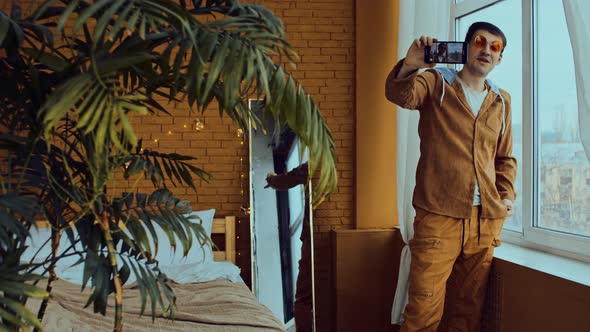 Young Man in Orange Glasses with Mobile Phone Standing Near Window